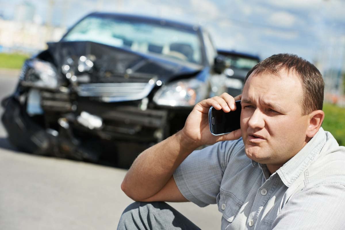 Homem falando ao celular e ao fundo um carro com a frente colidida 