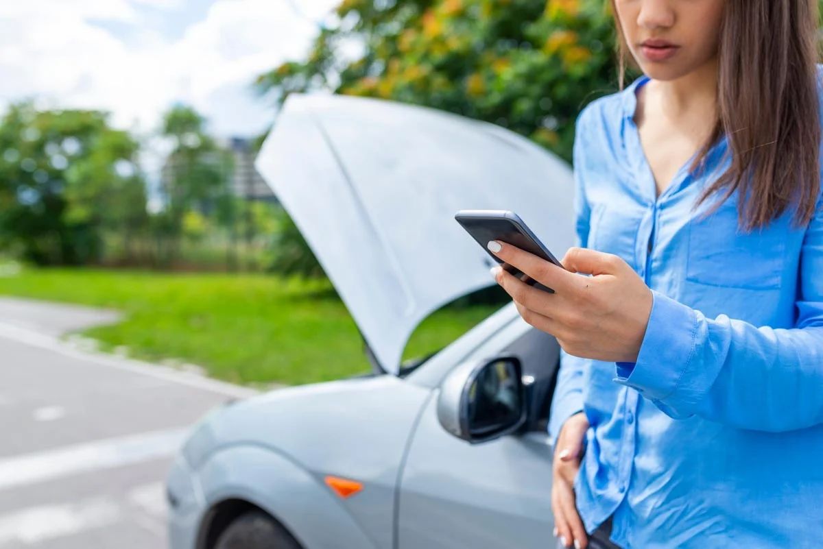 Mulher utlizando o celular para chamar resgate ao lado do carro quebrado  