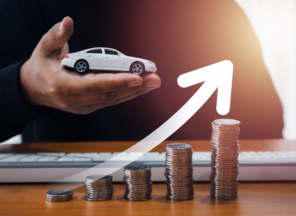 Moedas empilhadas e ao fundo um homem segurando uma miniatura de carro, representando o IPVA