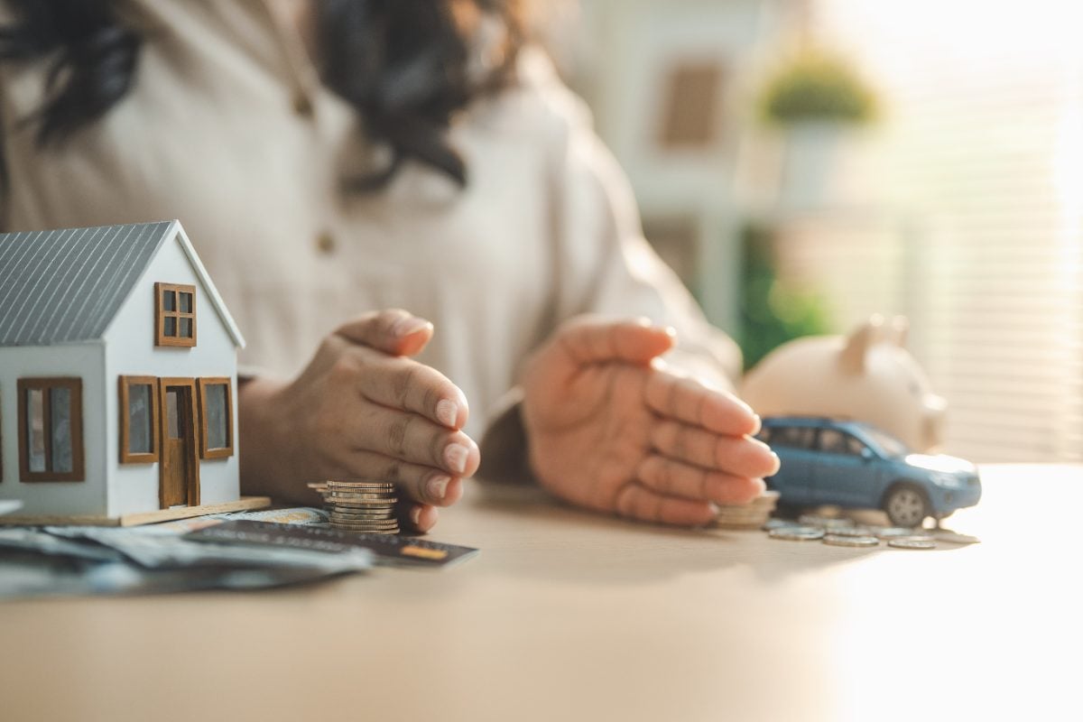 Mulher separando com as mãos maquete de casa, moedas e carro, representando o seguro auto 