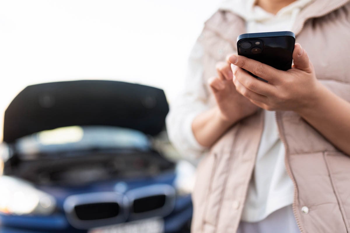 Pessoa ao telefone na frente de seu carro quebrado com o capô levantado 
