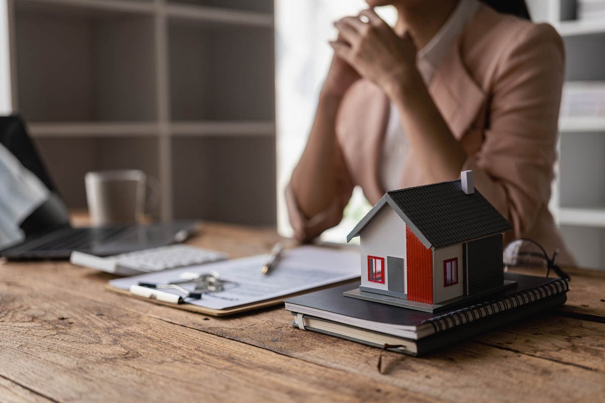 Miniatura de casa e caderno, papeis e notebook e uma pessoa sentada ao fundo