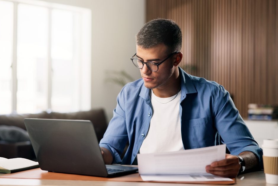 Homem usando notebook com papel nas mãos