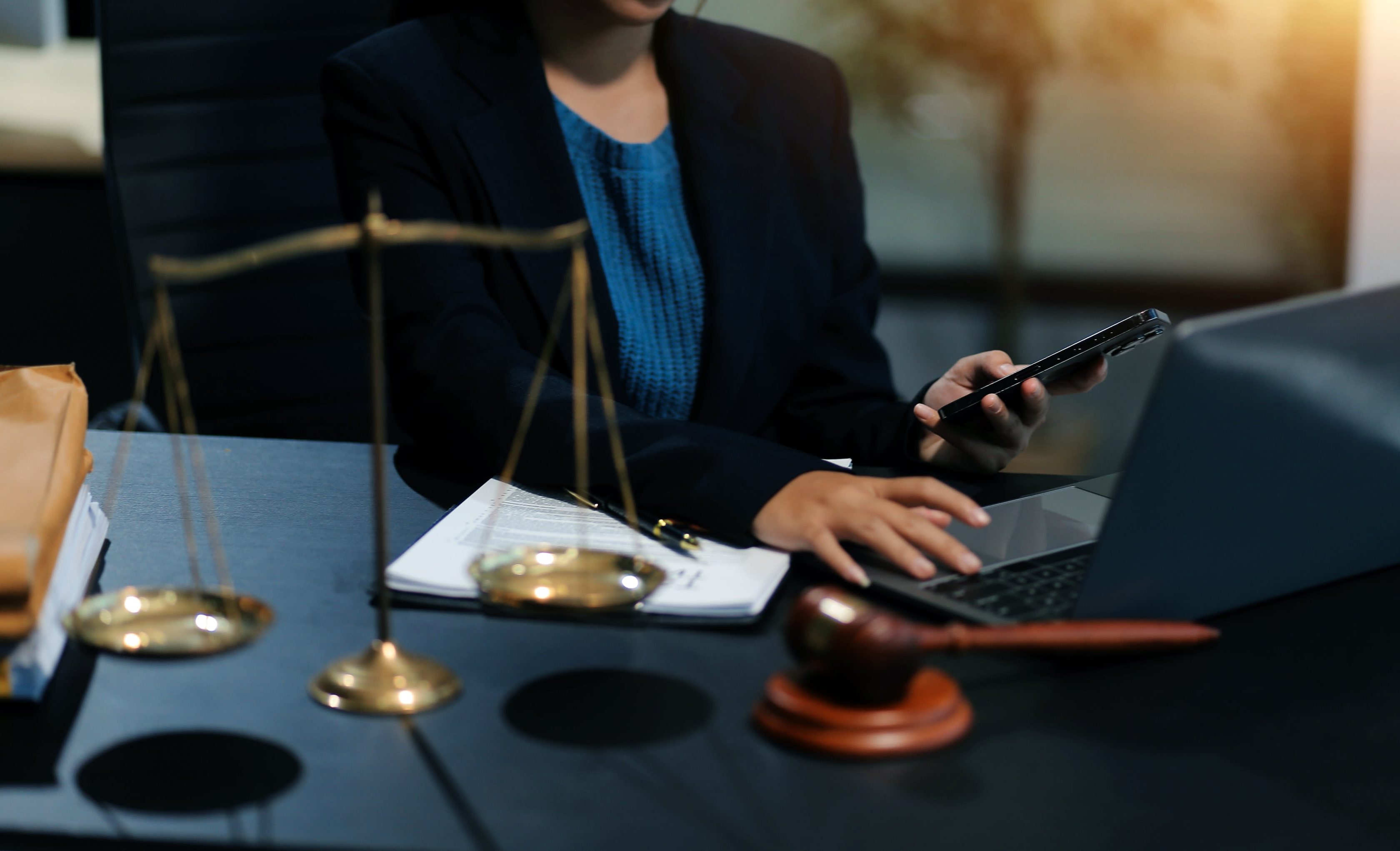 Profissional do direito trabalha com celular e laptop ao lado de balança e martelo da justiça.