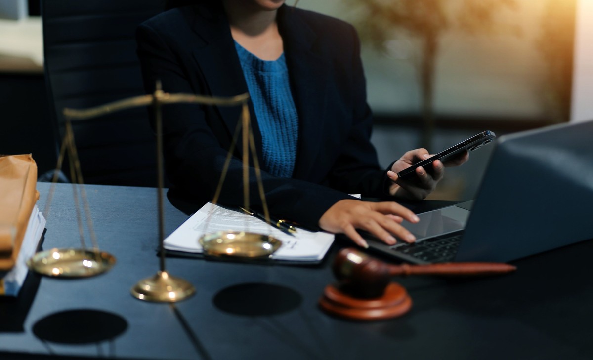 Profissional do direito trabalha com celular e laptop ao lado de balança e martelo da justiça.
