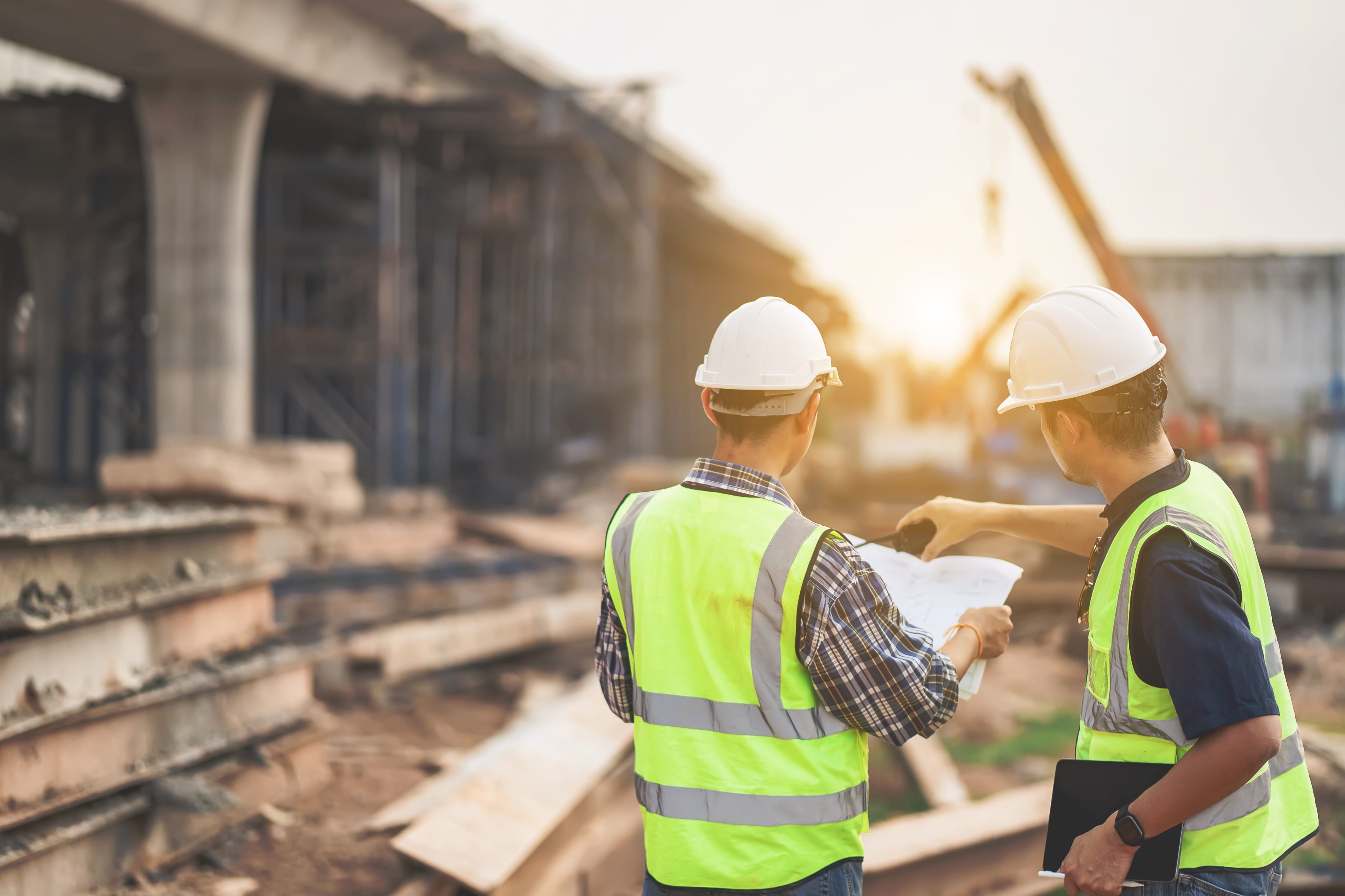 Dois  profissionais da construção civil em um canteiro de obras. Ambos vestem coletes de alta visibilidade e capacetes de segurança brancos. Um deles segura pranchas de papel, enquanto o outro aponta para algo no local. Ao fundo, é possível ver estruturas de concreto, andaimes, vigas e equipamentos pesados, como um guindaste.