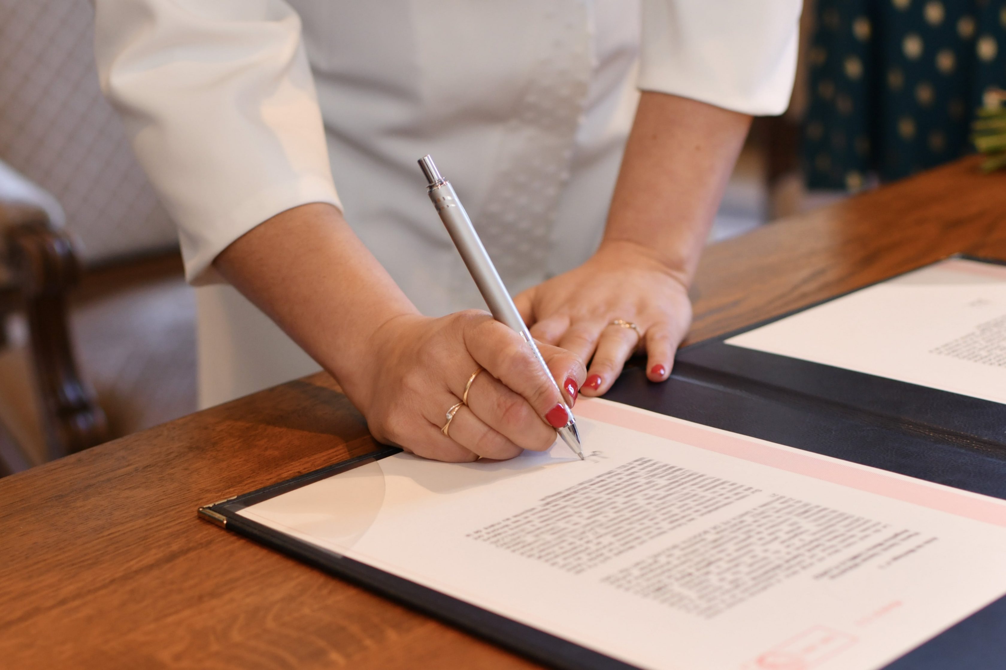 Mulher com a unha pintada de vermelho, está assinando um documento que está sobre uma mesa. Ela segura uma caneta, está vestida de branco e na imagem conseguimos visualizar somente os braços e as mãos.