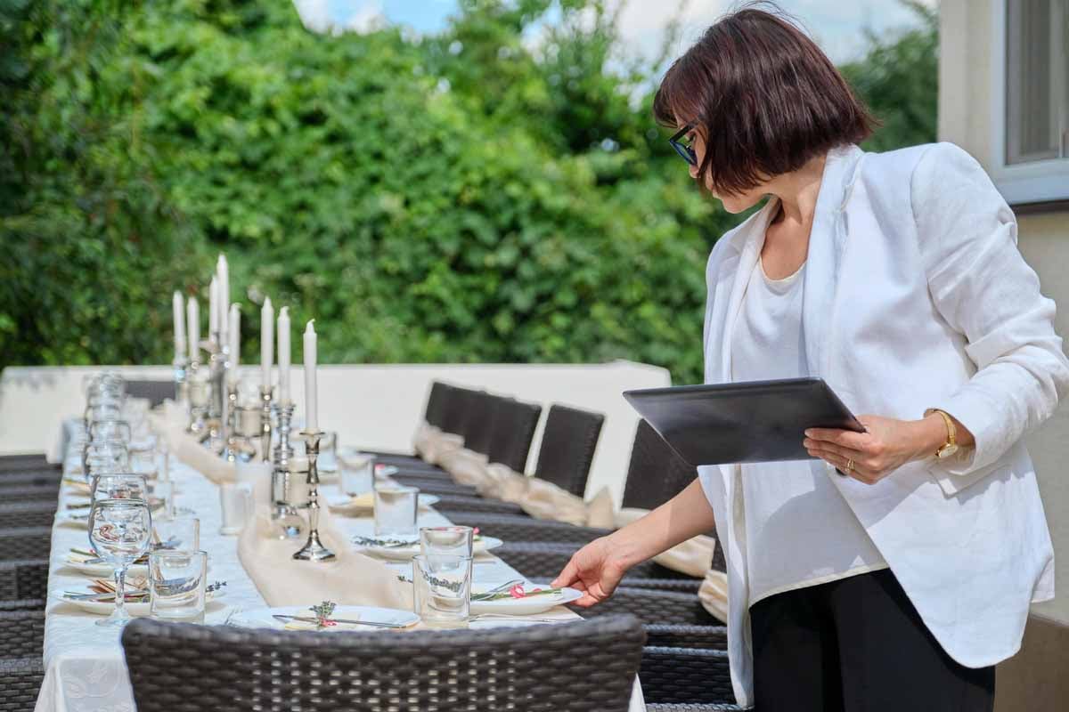 A imagem mostra uma mulher organizando uma mesa de jantar ao ar livre. A mulher está vestida com um blazer branco e segura um tablet, a mesa é longa  com castiçais altos com velas brancas. Taças de vinho e copos de água. Pratos organizados com guardanapos decorativos e ramos verdes, sugerindo atenção aos detalhes.