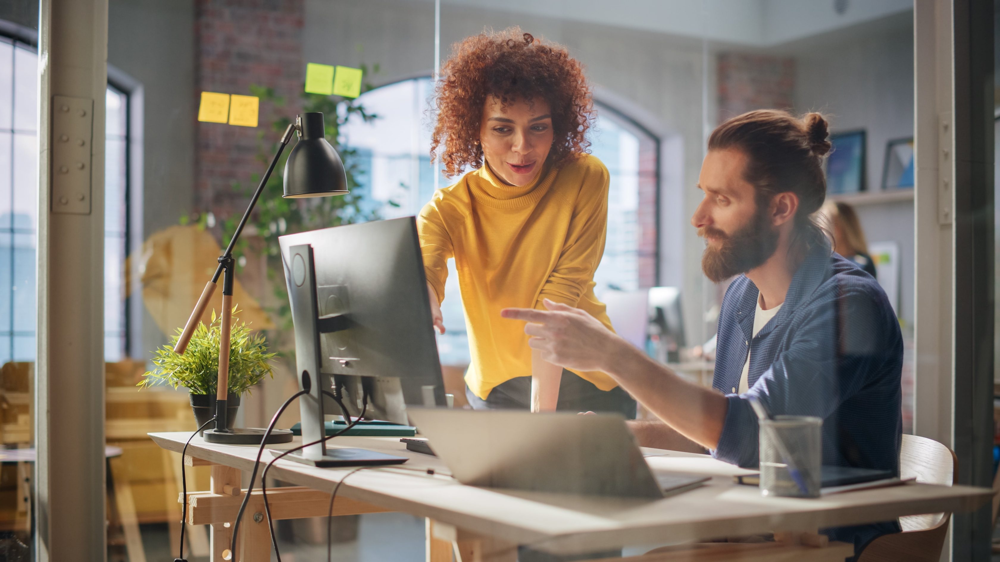 Dois profissionais em um ambiente de escritório. A mulher, vestindo um suéter amarelo, está em pé e inclinada para frente, observando a tela do computador com interesse. O homem, sentado à mesa, aponta para a tela. O ambiente  possui elementos como um vaso de planta, post-its coloridos no vidro e iluminação suave.