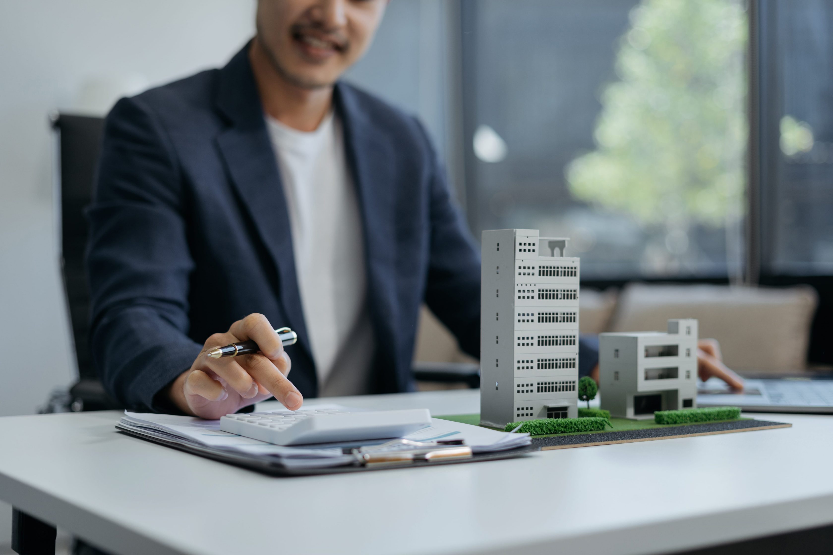 Homem em um escritório, está usando um blazer azul escuro e uma camiseta branca, usa uma calculadora com uma mão enquanto segura uma caneta com a outra. Sobre a mesa à sua frente, há duas maquetes de edifícios, é possível ver uma prancheta com documentos. Ao fundo, janelas amplas permitem a entrada de luz natural.