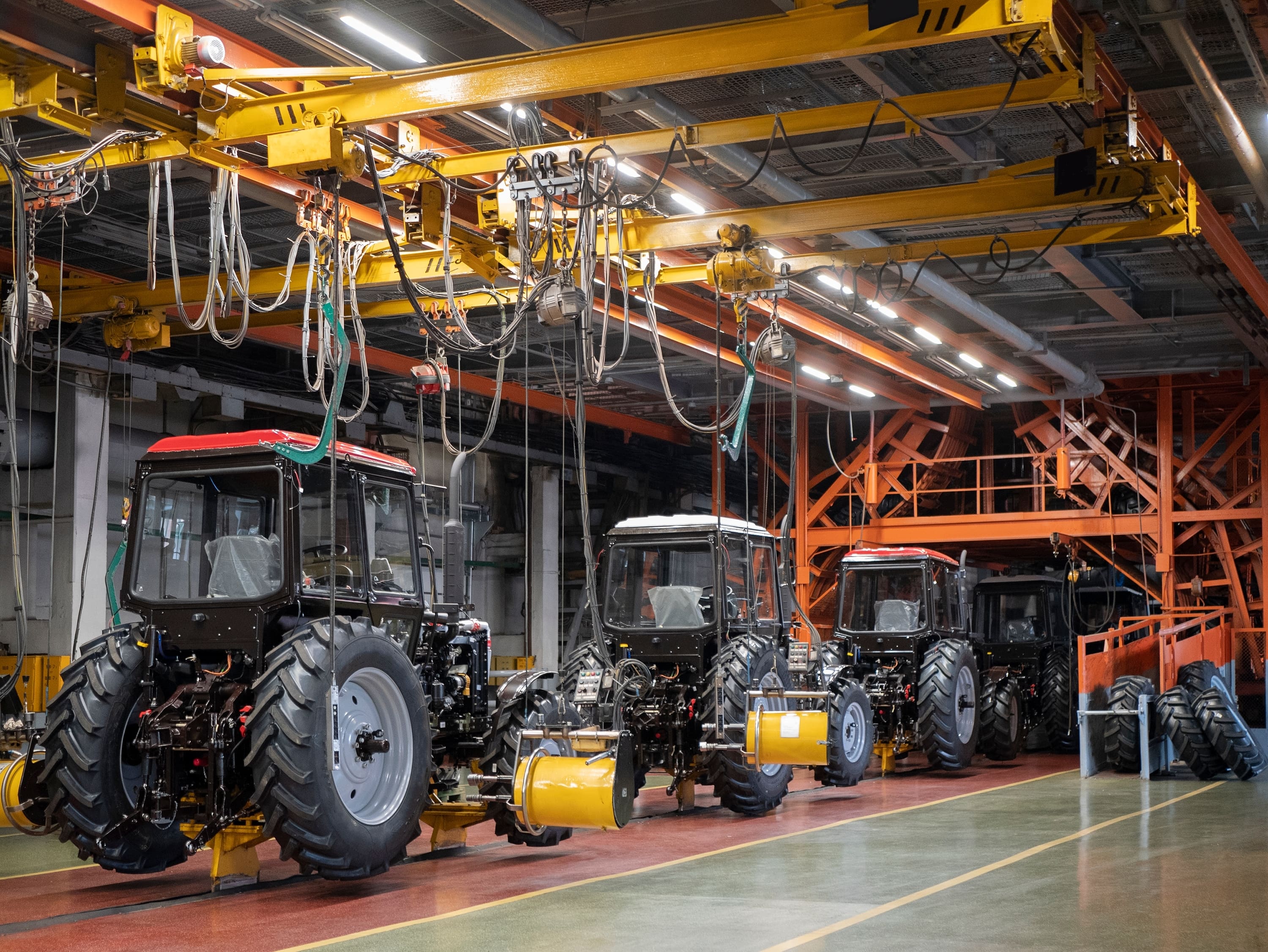 Linha de montagem industrial de tratores em uma fábrica. É possível ver vários tratores parcialmente montados enfileirados, com suas cabines e pneus traseiros já instalados. Acima da linha de montagem, há um sistema complexo de trilhos e cabos suspensos, provavelmente utilizado para movimentação de peças e ferramentas. A estrutura metálica da fábrica, com cores predominantes em amarelo e laranja, reforça o ambiente industrial e automatizado do local. A iluminação é forte e direcionada, permitindo visibilidade clara para os trabalhos de montagem..
