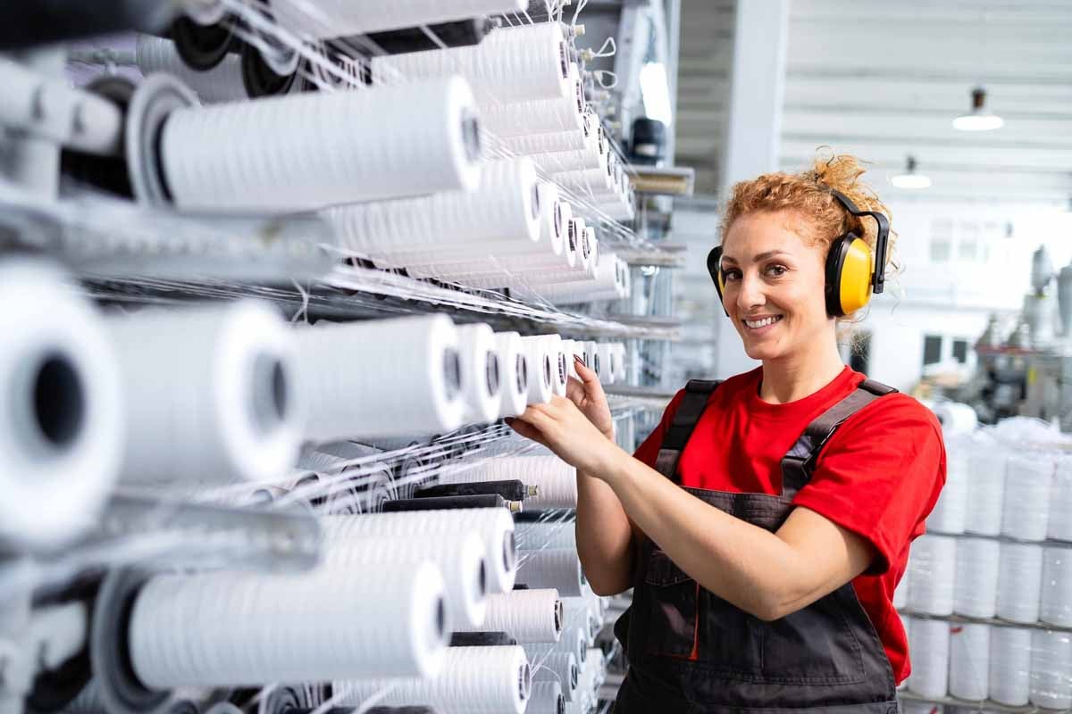 Uma mulher trabalhando em uma fábrica têxtil, operando rolos de fio branco alinhados em prateleiras metálicas. A mulher veste uniforme com camiseta vermelha e macacão preto, usa protetores auriculares amarelos. Ela sorri enquanto realiza sua atividade. Ao fundo, é possível ver mais equipamentos e materiais da indústria.