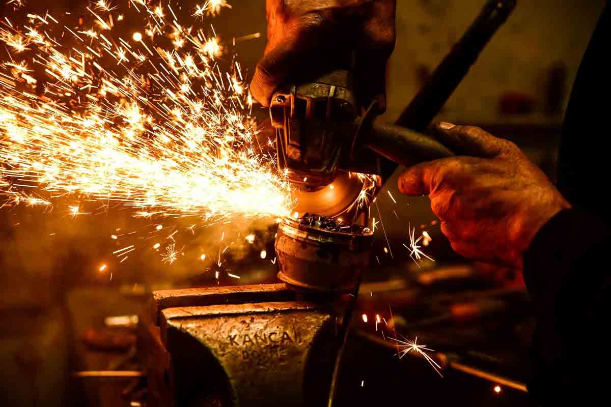 Um trabalhador utilizando uma esmerilhadeira para cortar uma peça metálica presa em um torno de bancada, uma intensa emissão de faíscas incandescentes.