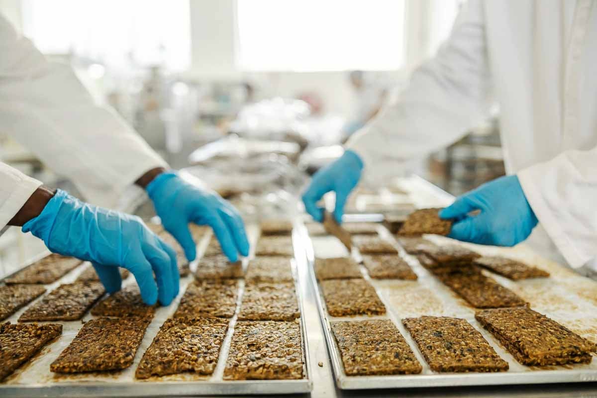 Técnicos manuseiam barras de cereais em linha de produção com rigoroso controle de qualidade.