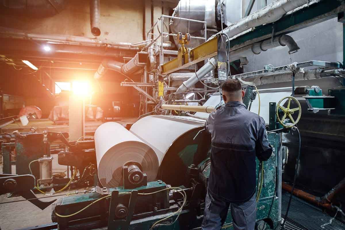 Interior de uma fábrica de papel durante o processo de produção. Um operador, de costas para a câmera e vestindo uniforme industrial, está monitorando uma grande máquina que transforma celulose em rolos contínuos de papel. O ambiente industrial é dominado por estruturas metálicas, tubulações, cilindros e equipamentos robustos. No centro da cena, grandes bobinas de papel já parcialmente enroladas são visíveis. Ao fundo, a luz solar entra pela janela, criando um contraste com a iluminação artificial do local, conferindo um toque dramático à cena.