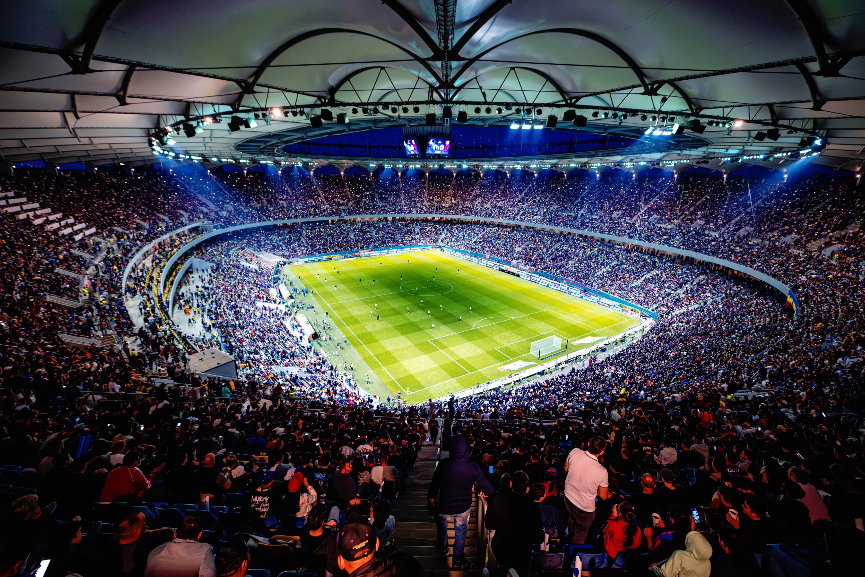 Estádio de futebol lotado a noite. A vista é panorâmica, capturada a partir das arquibancadas superiores que estão completamente lotadas por. O campo está iluminado por potentes refletores.