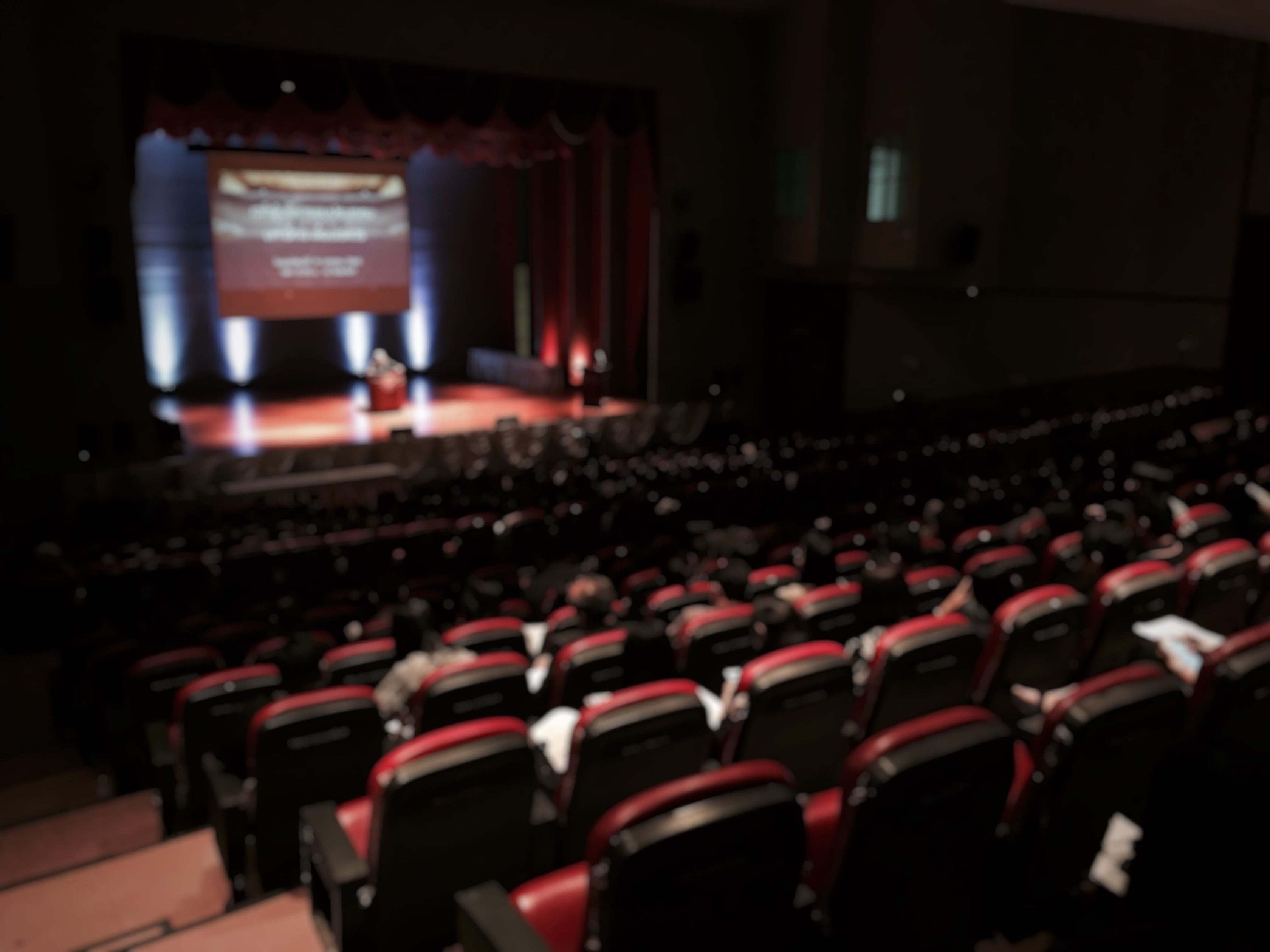 A imagem mostra o interior de um auditório com cadeiras vermelhas e pretas, onde ocorre um evento formal. A imagem está desfocada, mas é possível identificar uma pessoa em pé no palco, próxima a um púlpito, com uma projeção ao fundo e iluminação voltada para a área central. O auditório está relativamente.