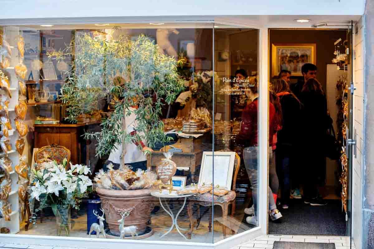 A imagem mostra a fachada de uma loja. A loja está bem iluminada e apresenta uma vitrine repleta de itens decorativos, flores, embalagens de produtos artesanais e uma árvore ornamental. Do lado direito da imagem, é possível ver várias pessoas dentro da loja. A decoração com móveis antigos, objetos dourados e detalhes natalinos como cervos decorativos.
