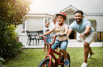 Mãe e filha sorrindo e observando o pai ensinando o filho a andar de bicicleta.