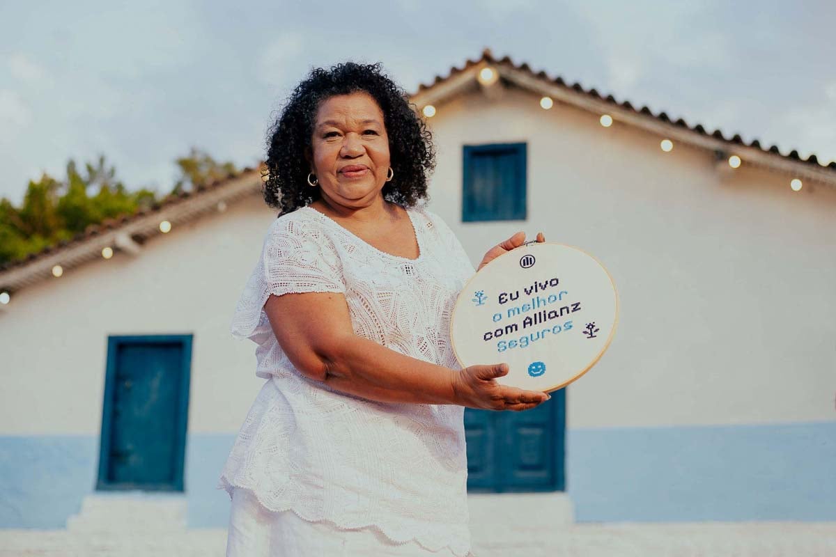 Mulher segurando placa da campanha “Vivendo o Melhor”, da Allianz Seguros, em frente a uma casa, representando histórias reais de cuidado e proteção.