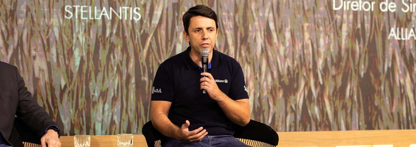 Diretor de Sinistro Auto da Allianz fala ao microfone durante painel, sentado em cadeira com fundo de palco iluminado.