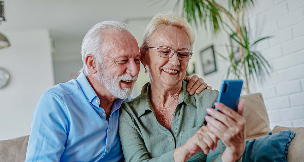 Imagem de um casal de idosos sorrindo e utilizando o celular