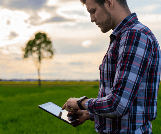 Homem utilizando um tablet no meio do campo com uma árvore ao fundo