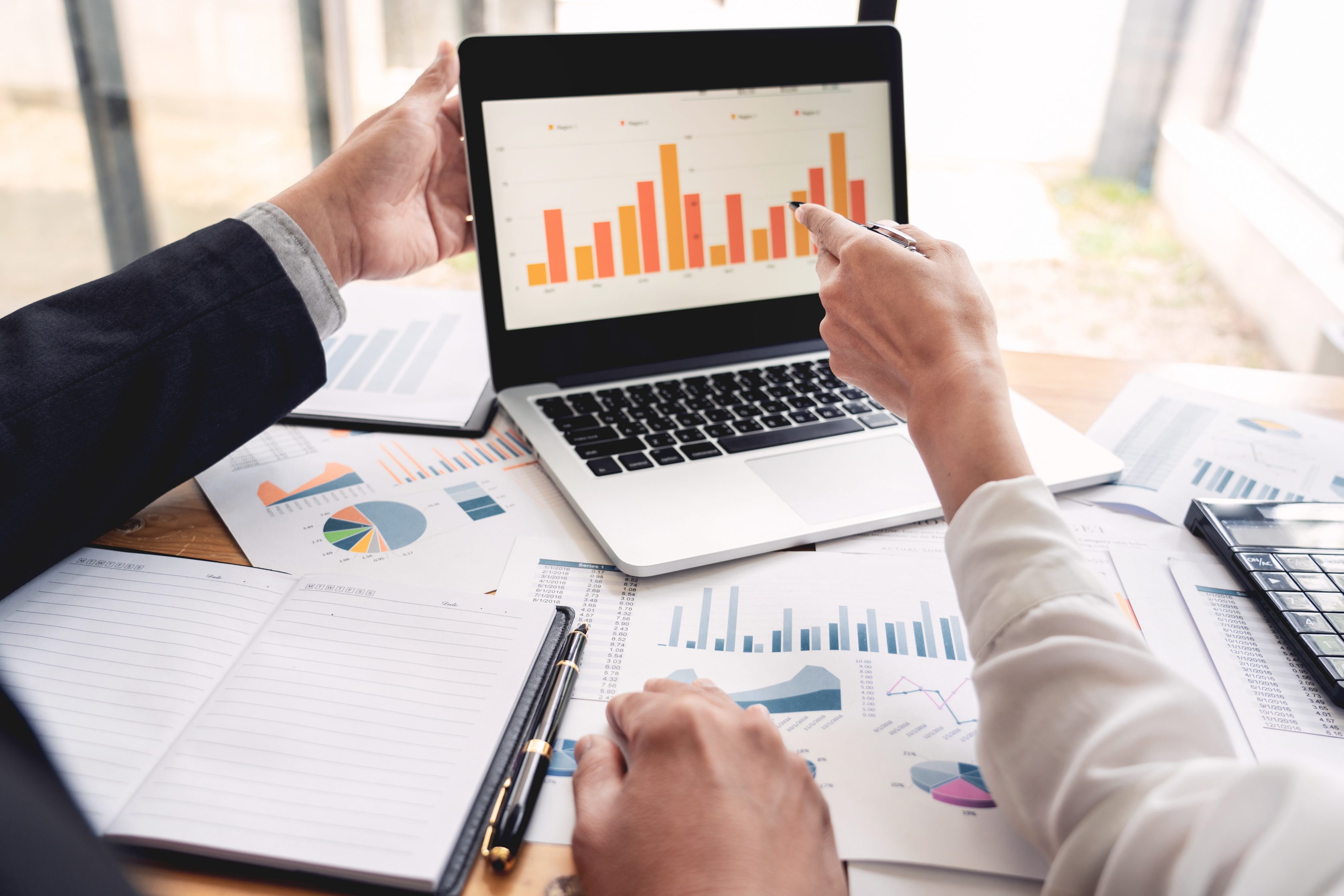 Pessoas analisando um computador com um dashboard e papéis com dados