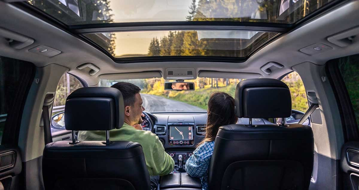 Homem dirigindo seu carro em área rural tranquilamente, ao lado de sua esposa. 