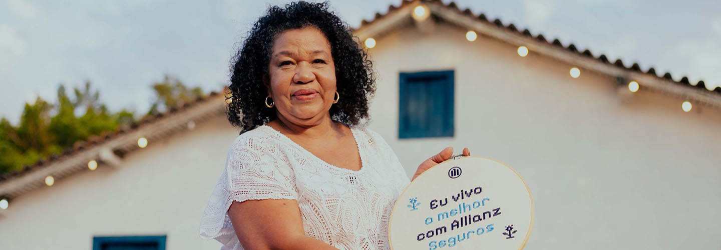 Mulher segurando placa da campanha “Vivendo o Melhor”, da Allianz Seguros, em frente a uma casa, representando histórias reais de cuidado e proteção.