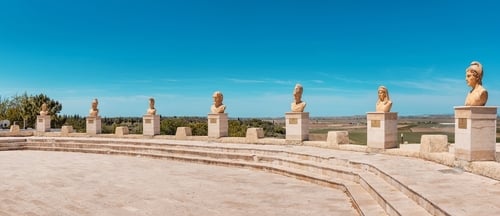 Bustos clássicos alinhados em área externa com vista panorâmica, remetendo à arte e à mitologia grega
