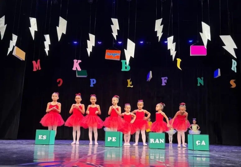 Crianças no palco durante apresentação de ballet, usando figurinos vermelhos, em cenário lúdico que integra espetáculo cultural promovido pela ABA.